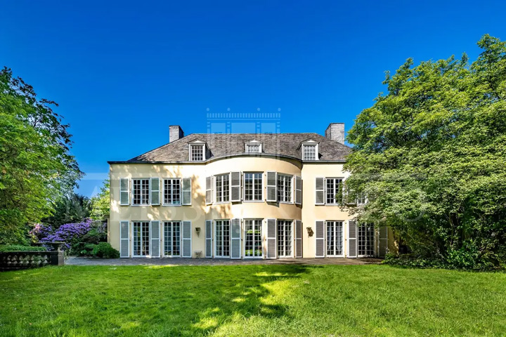 Eine majestätische, klassizistische Villa in französischem Château-Stil mit eleganter, cremefarbener Fassade und traditionellem grauen Schieferdach. Das zweistöckige Herrenhaus zeigt symmetrische Architektur mit großen, bodentiefen Fenstern und charakteristischen hellblauen Fensterläden an der gesamten Fassade. Das Gebäude verfügt über mehrere Dachgauben mit kleinen Fenstern im Mansarddach und zwei Schornsteine. Die repräsentative Gartenseite des Anwesens zeigt eine breite Rasenfläche in perfektem Zustand, die sich großzügig vor dem Gebäude erstreckt. Das Anwesen ist von einem parkähnlichen Garten mit alten, ausladenden Laubbäumen umgeben, die natürlichen Rahmen und Privatsphäre schaffen.