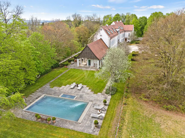 Luftaufnahme eines luxuriösen Anwesens mit modernem Wohnhaus und exklusiver Poolanlage in weitläufiger Parklandschaft. Das zweistöckige Hauptgebäude zeigt zeitgenössische Architektur mit weißer Fassade, großzügigen Glasfronten und roten Ziegeldächern. Der Höhepunkt der Außenanlage ist ein großer, rechteckiger Swimmingpool mit türkisblauem Wasser, der von einer eleganten Terrasse aus hellem Naturstein umgeben ist.