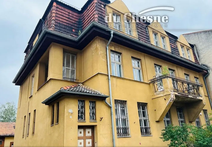 Ein mehrstöckiges Gebäude aus gelbem Backstein mit charakteristischen roten Ziegeldächern und weißen Sprossenfenstern. Die Fassade zeigt klassische Gründerzeit-Architektur mit verschiedenen Gebäudeteilen und einem kleinen Balkon mit schwarzem Eisengeländer. Im unteren Bereich sind Fenster mit Gittern zu sehen. Das Gebäude wird von grüner Vegetation umgeben.