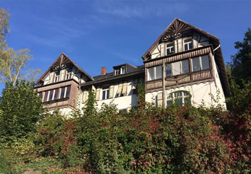 Eine Villa mit charakteristischen Fachwerk-Elementen. Das mehrstöckige Gebäude besteht aus mehreren verbundenen Gebäudeteilen mit unterschiedlichen Dachformen und roten Ziegeldächern. Die Fassade kombiniert weiße Putzwände mit dunklen Holzbalken im traditionellen Fachwerk-Stil. Markante architektonische Details sind die reich verzierten Holzbalkone mit geschnitzten Geländern, große Sprossenfenster und verschiedene Dacherker und Gauben.
