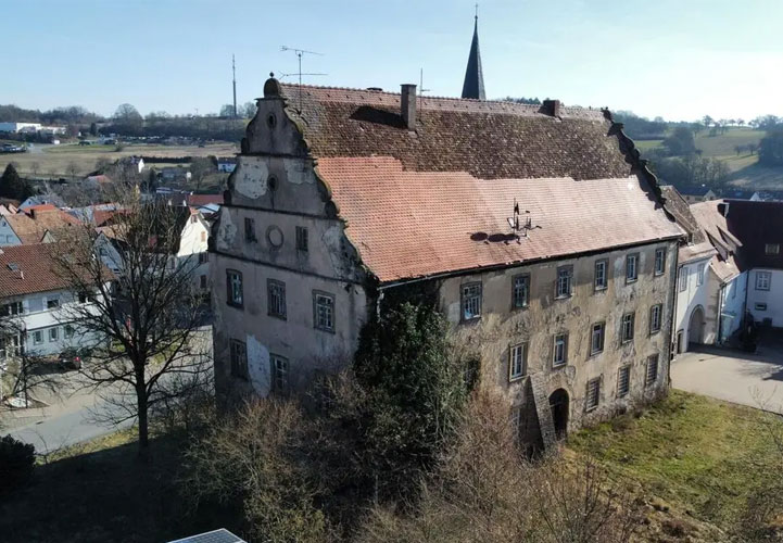 Luftaufnahme eines historischen Schlosses in ländlicher Umgebung. Das große, mehrstöckige Hauptgebäude hat charakteristische rote Ziegeldächer und helle Fassaden mit regelmäßig angeordneten Fenstern. Links erhebt sich ein massiver, eckiger Turm mit grauer Steinfassade und mehreren Geschossen. Der Komplex liegt inmitten einer kleinen Ortschaft mit traditionellen Häusern mit roten Dächern.