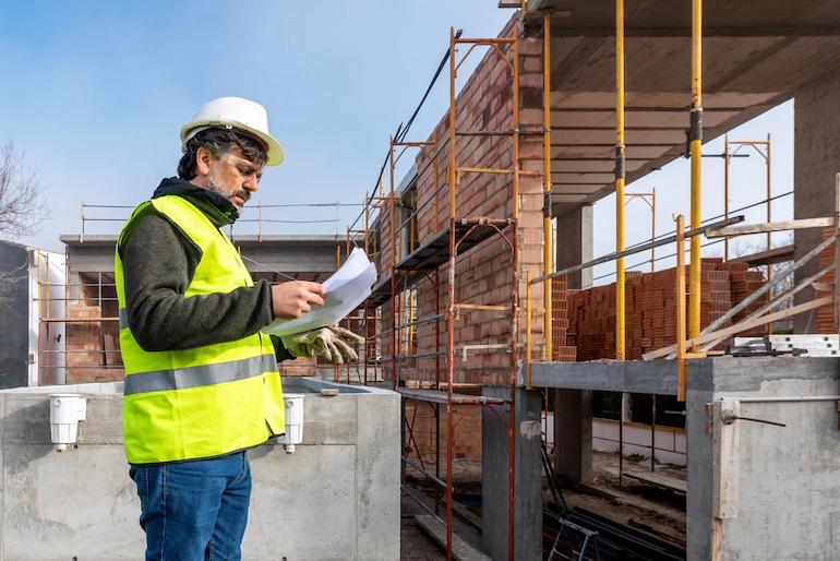 Mann mit Warnweste und Helm hat Klemmbrett in der Hand und steht vor einem Gebäude das saniert wird