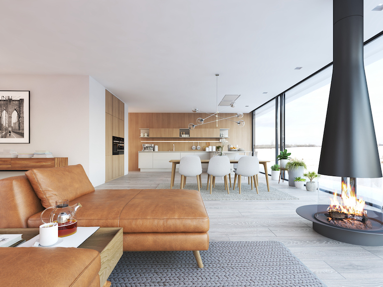 Ein helles Wohn- und Esszimmer mit Holzvertäfelung und weißen Stühlen und einem Kamin,  sowie einer Fensterfront rechts, das Sofa und der Kamin sind voll abgebildet