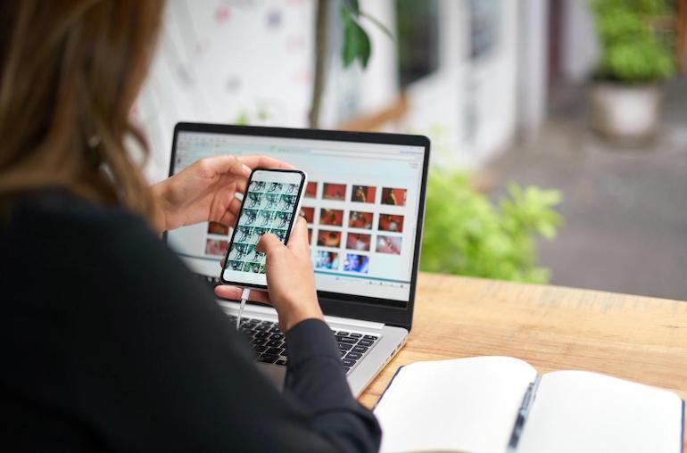 Vermieterin scrollt am Smartphone geschossene Bilder ihrer Immobilie durch, die sie am Laptop vor sich bearbeiten möchte.  Neben ihr liegt ein Notizheft.