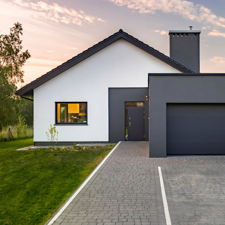 Weißes Einfamilienhaus aus Sicht der Einfahrt mit grünem Rasen und beginnendem Sonnenuntergang im Hintergrund