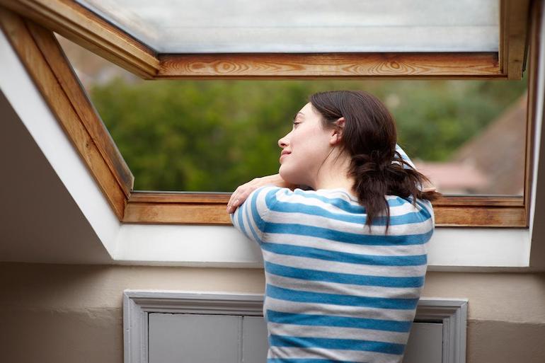 Frau lehnt sich lächelnd aus dem Dachfenster und genießt das Grün und den Sommerwind